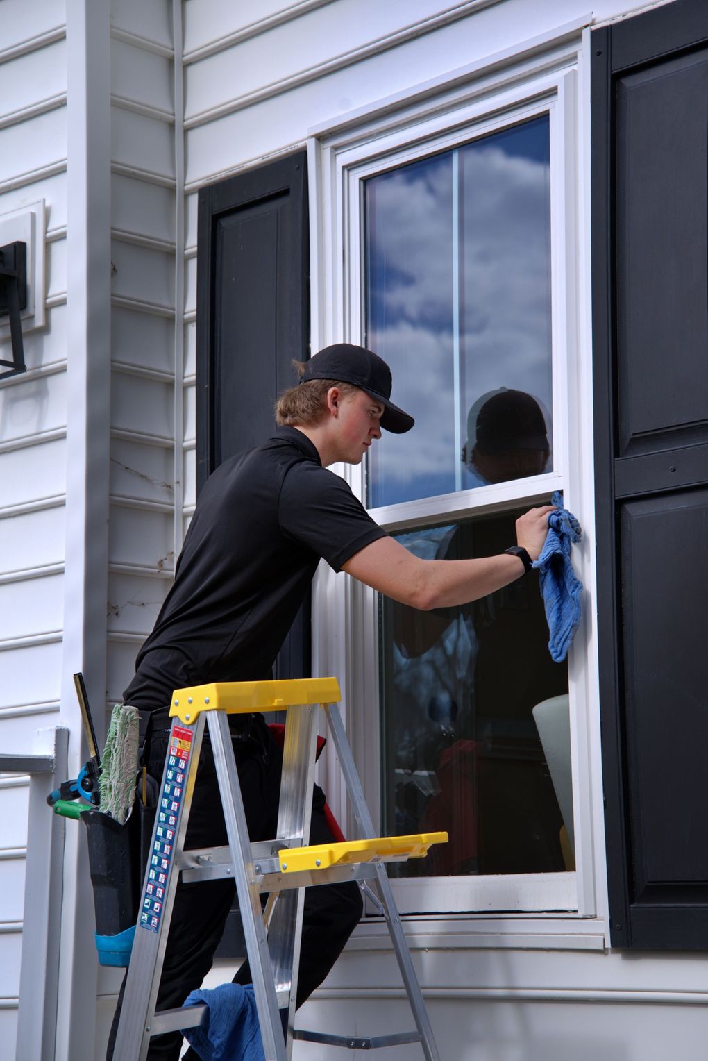 Person cleaning a window on a house, using a ladder and blue cloth.