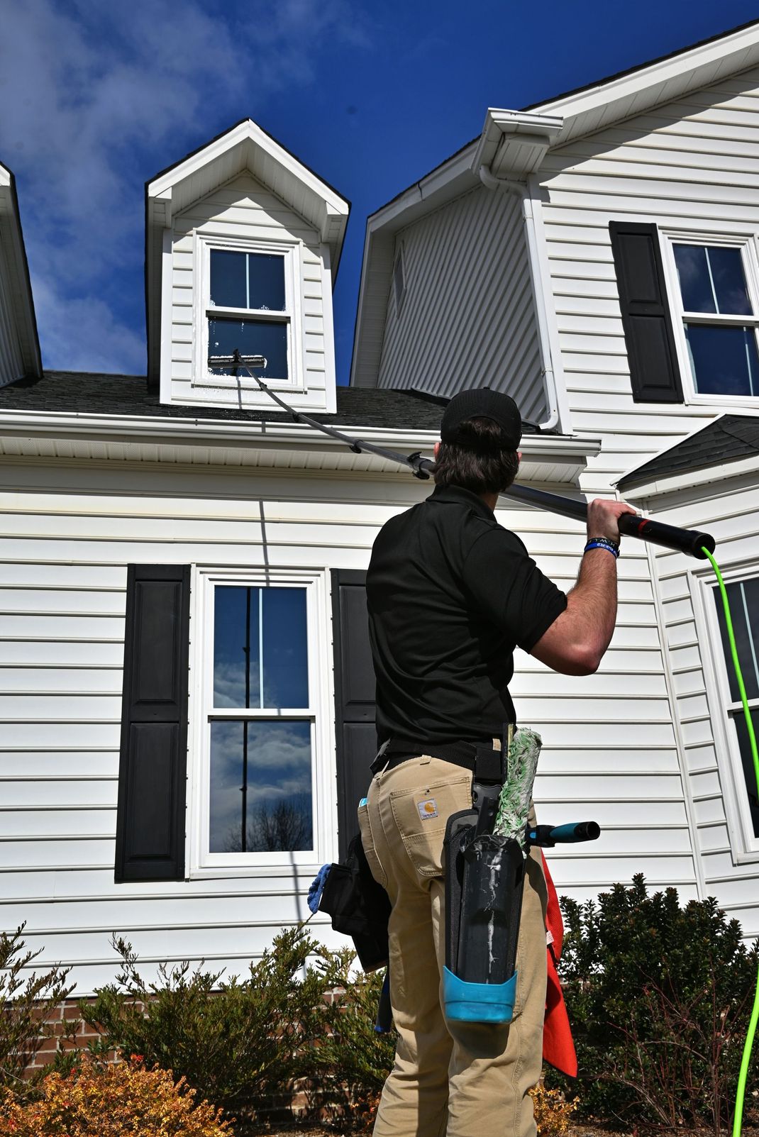 Person cleaning windows on a two-story house. He is standing on a small roof and has cleaning supplies.