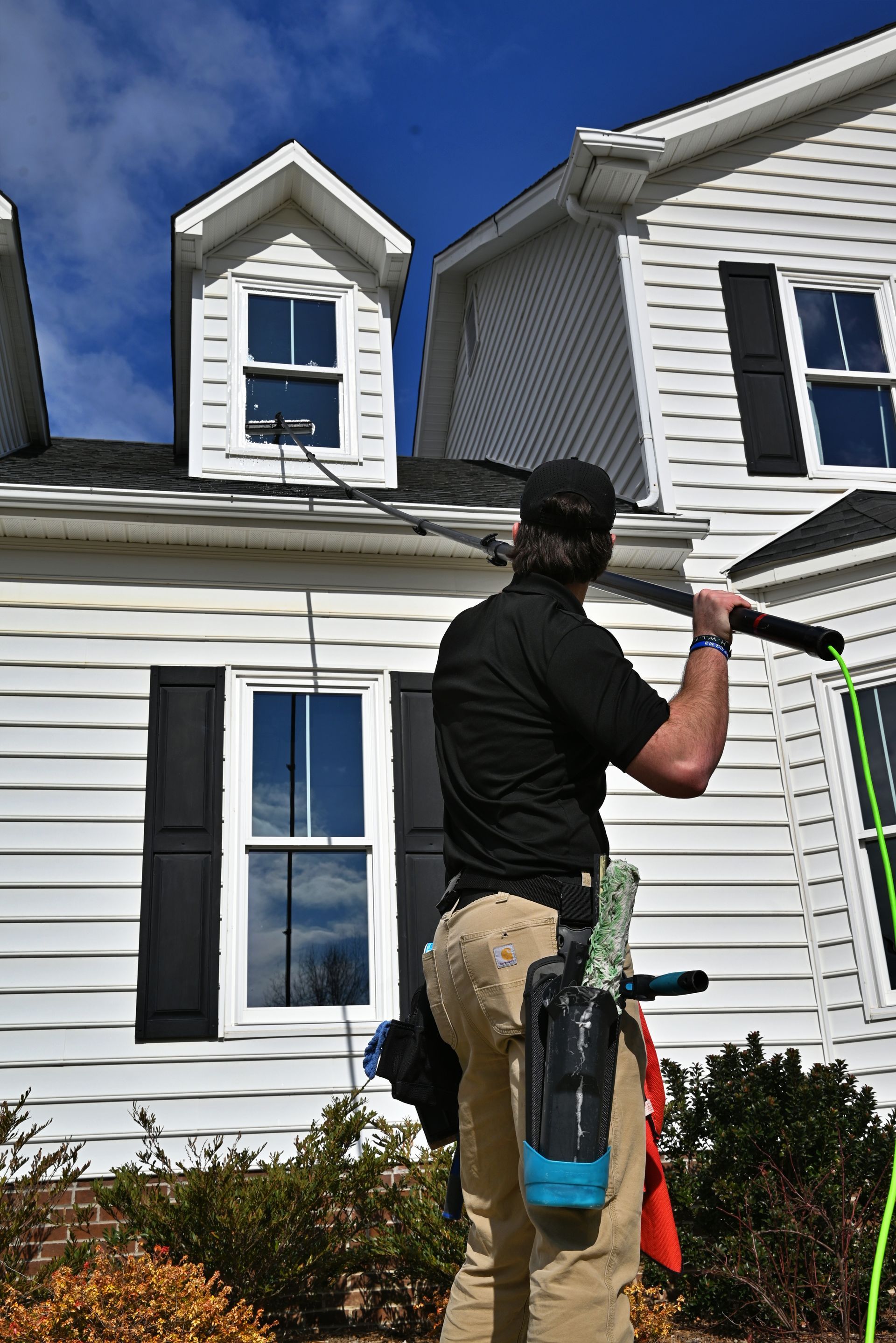 Person cleaning windows on a two-story house. He is standing on a small roof and has cleaning supplies.