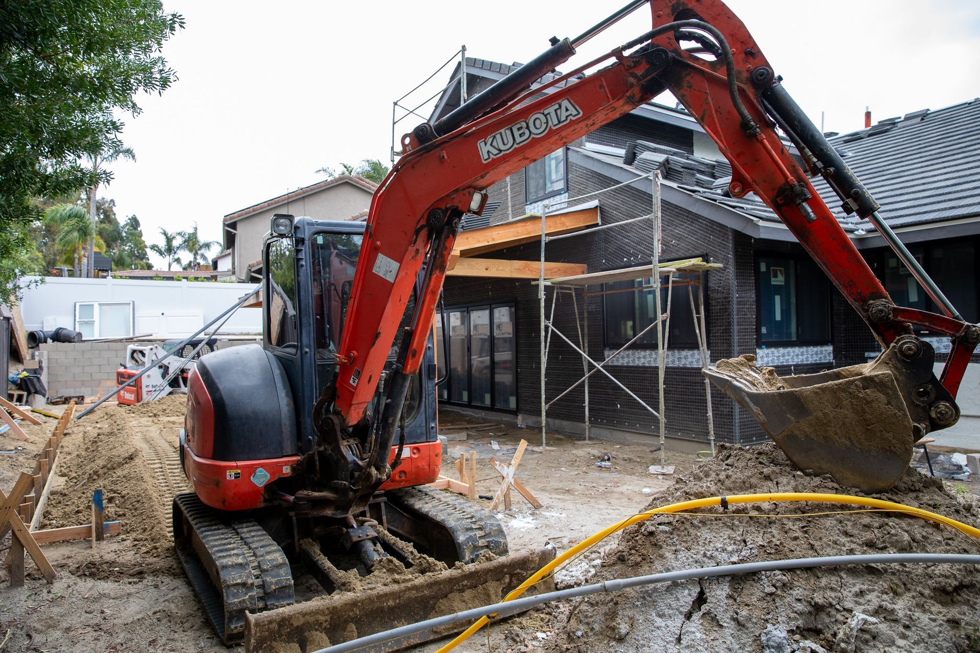 A red and black excavator is digging a hole in front of a house under construction.