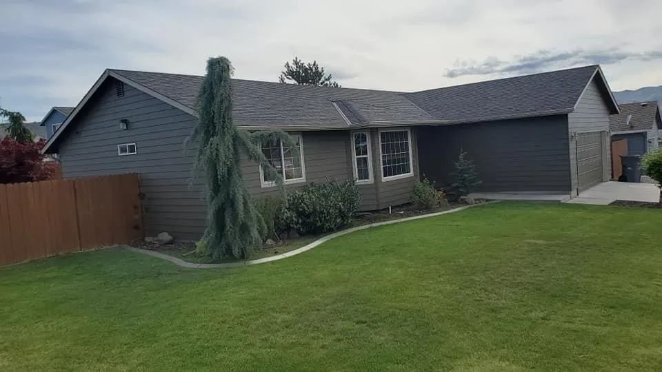 House with gray siding and brown roof, green lawn, and tall, thin evergreen tree.