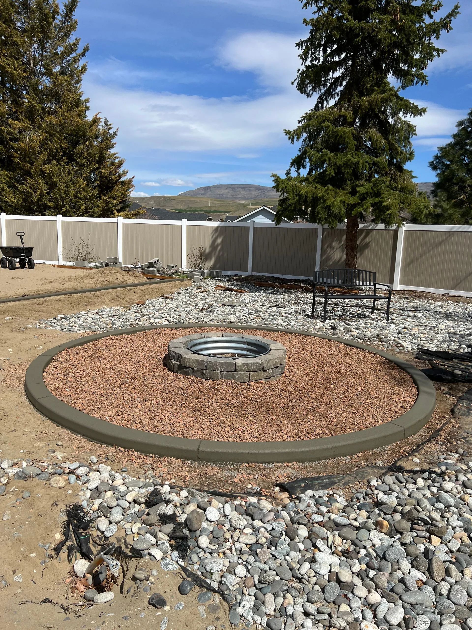 A newly constructed circular fire pit with brown rocks, surrounded by concrete border, in a backyard.