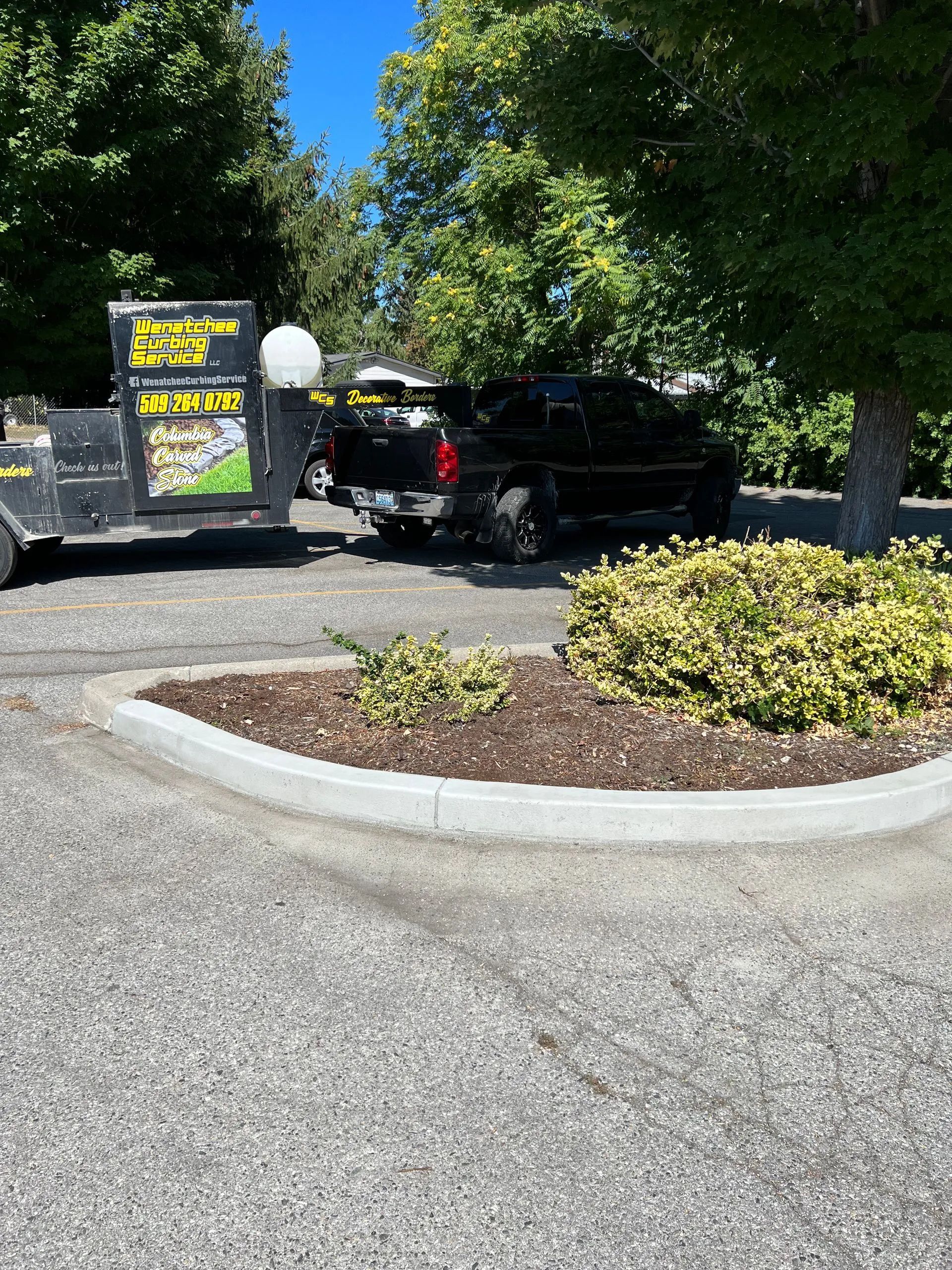 Black truck towing trailer parked in a gravel lot next to a landscaped area and trees.