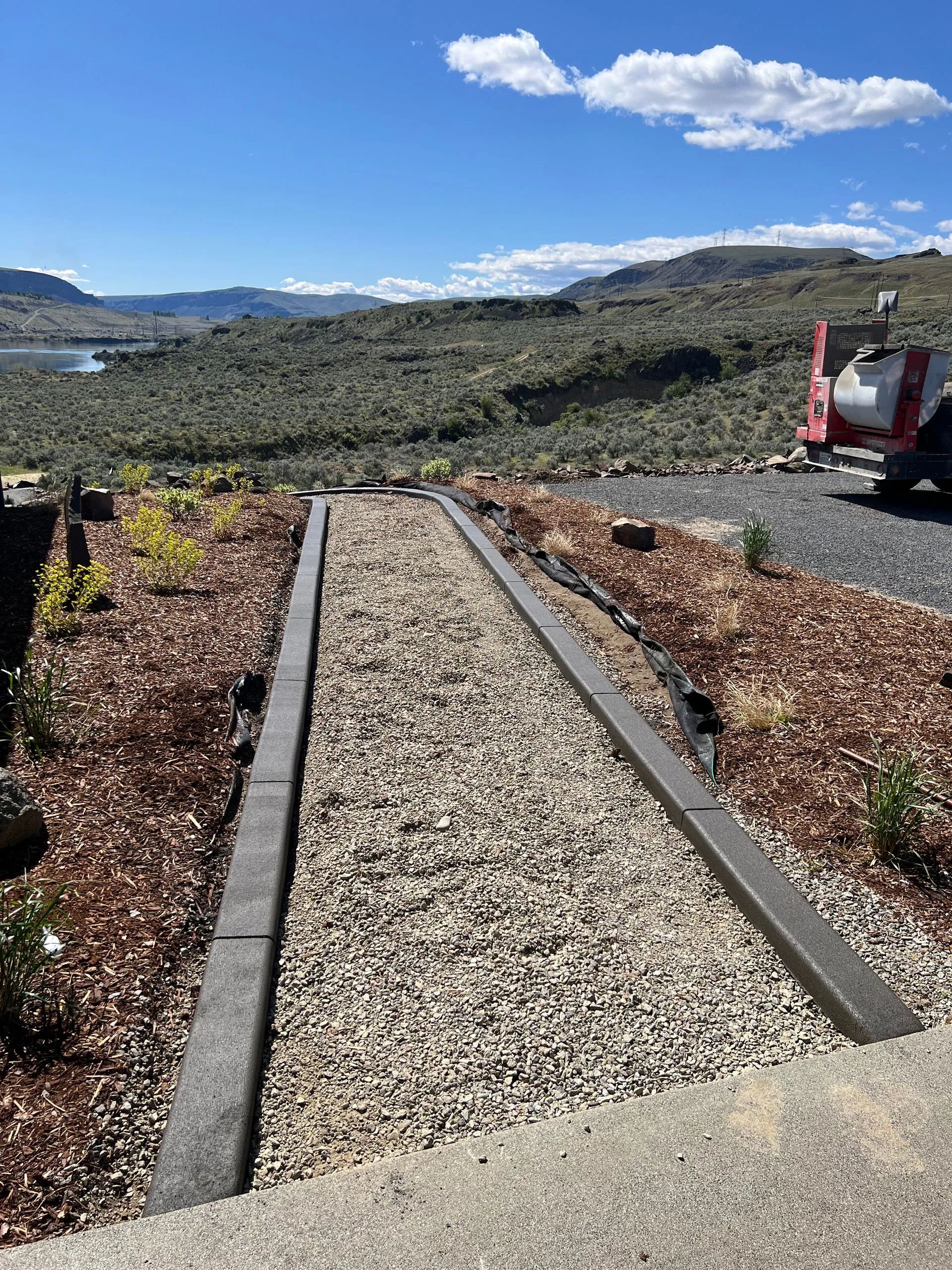 A gravel path with gray borders leads through a landscape of plants and brown mulch. Blue sky and hills in background.
