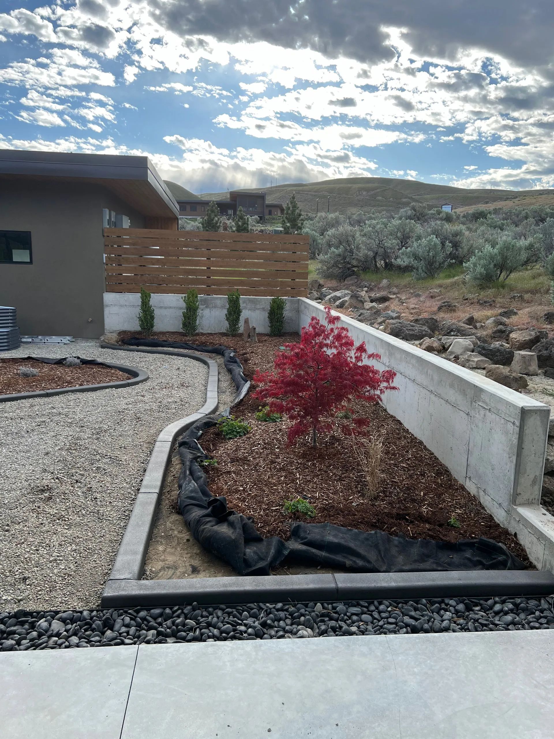 Landscaped yard with stone borders, red maple tree, wood fence, and hillside background.