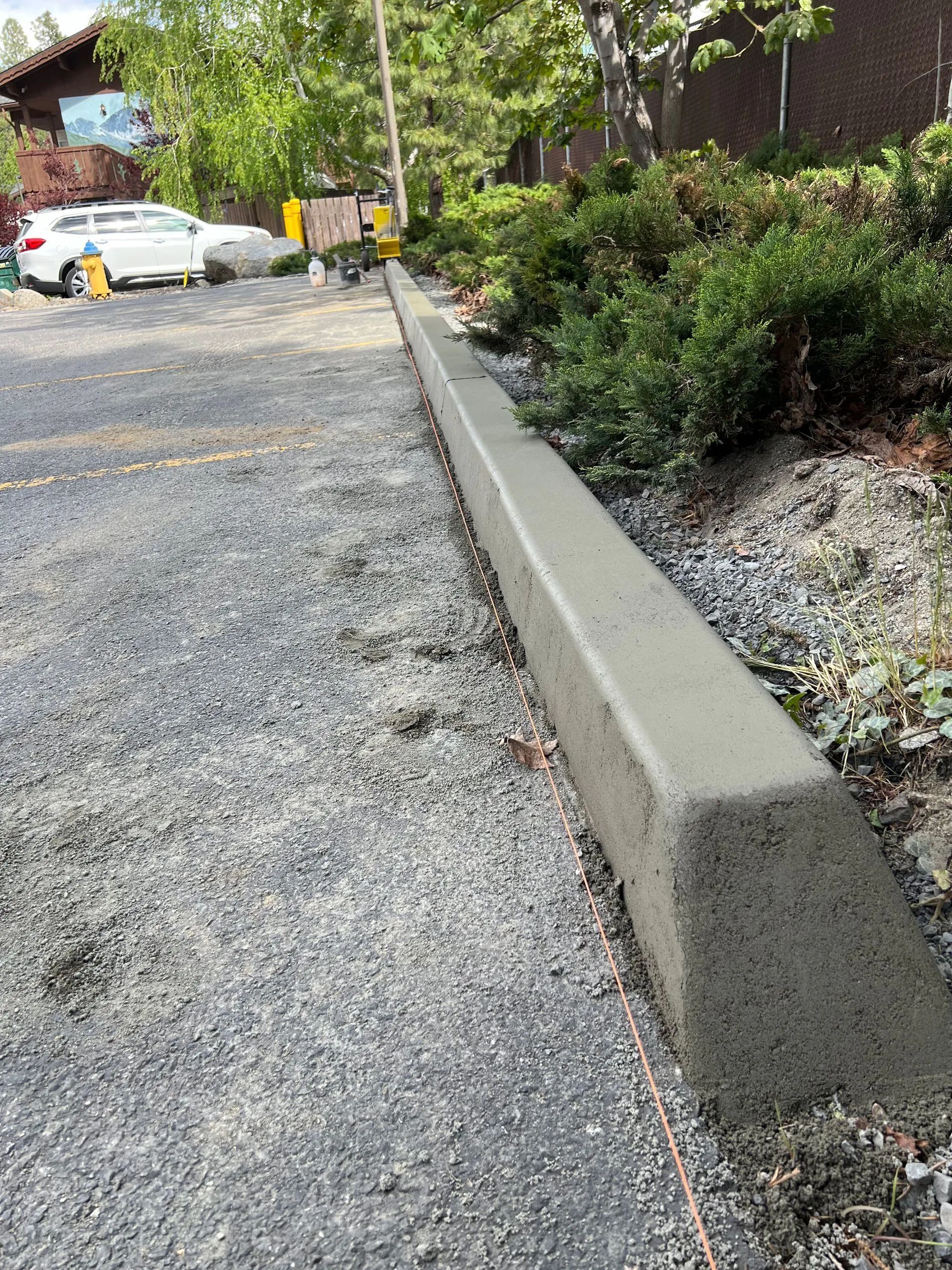Newly poured concrete curb along a gravel driveway; trees and parked white car in the background.