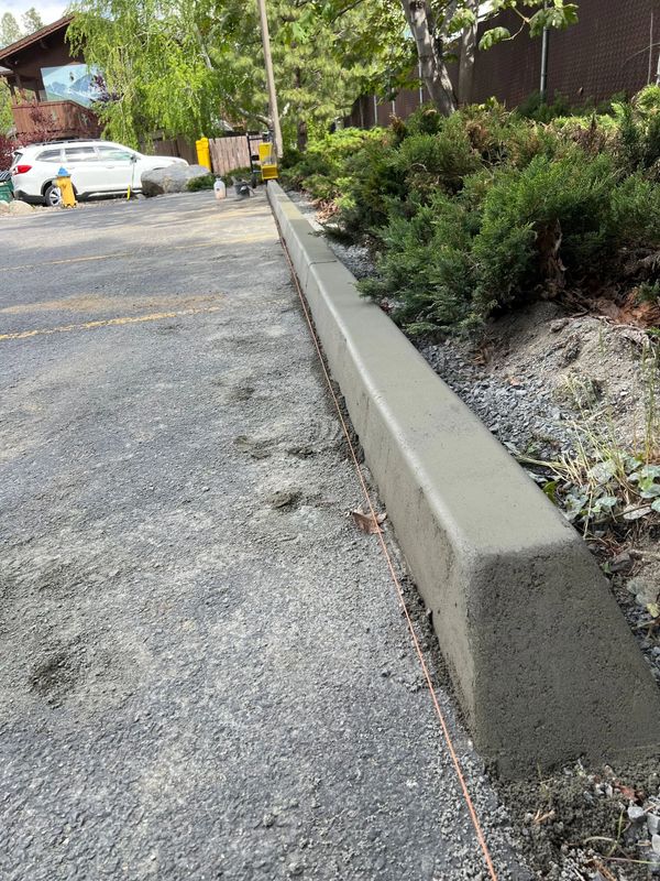 Newly poured concrete curb along a gravel road, with vegetation and buildings in the background.