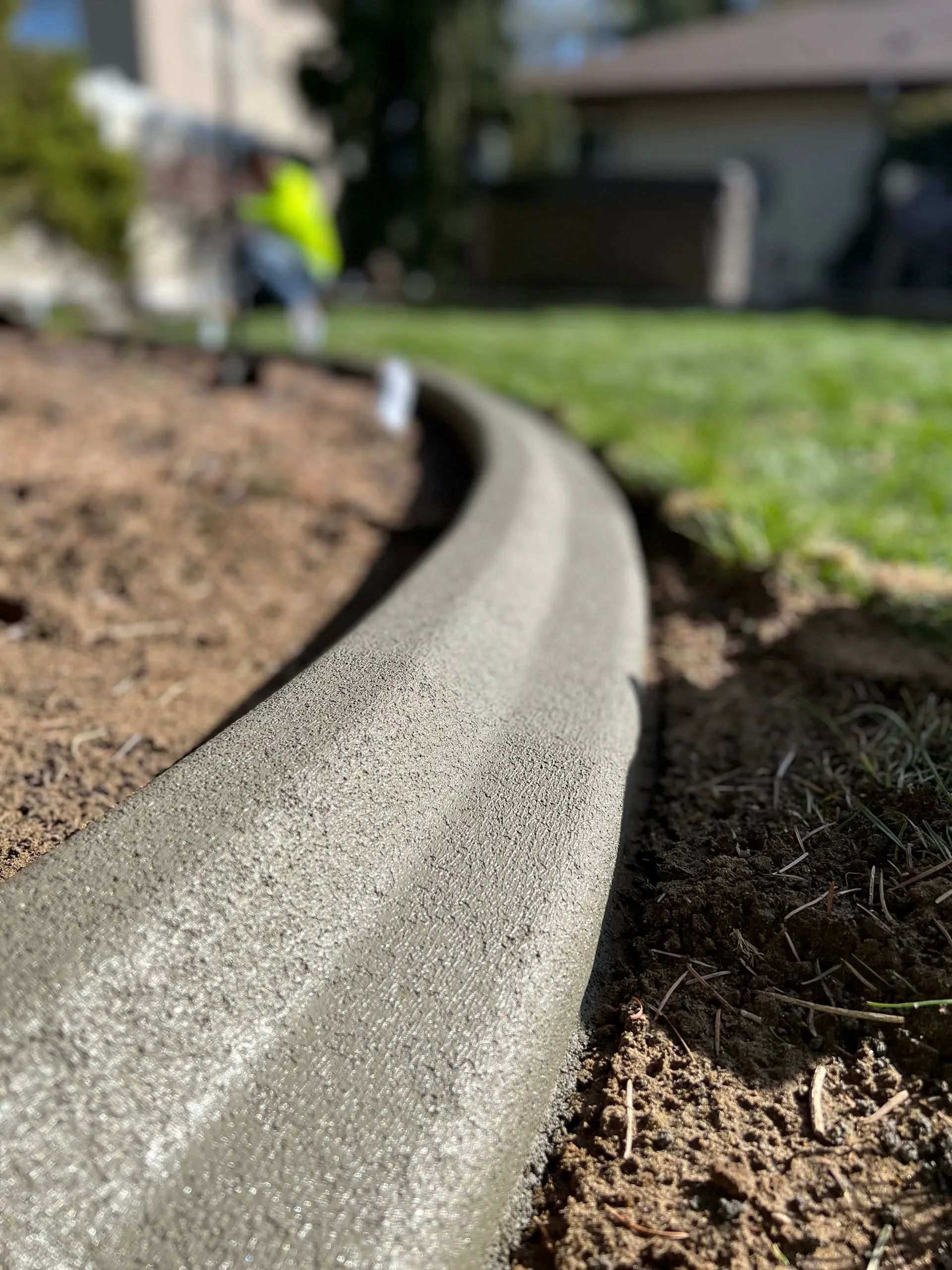 Newly poured concrete curbing bordering a lawn and garden bed, worker in background.