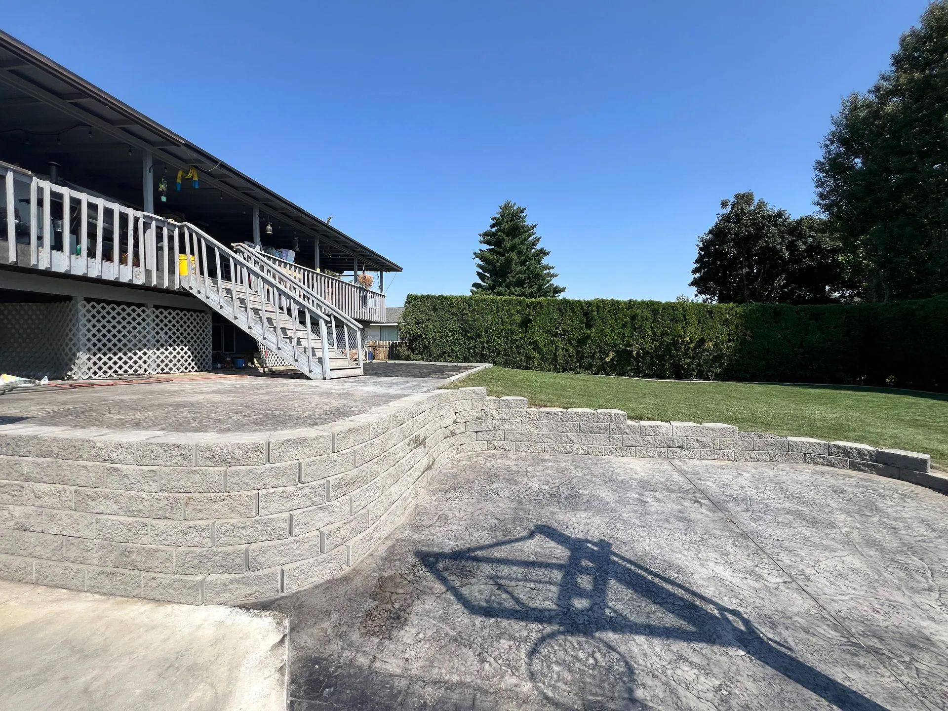 Backyard with stone retaining wall, basketball hoop shadow, and house with wooden stairs under a blue sky.