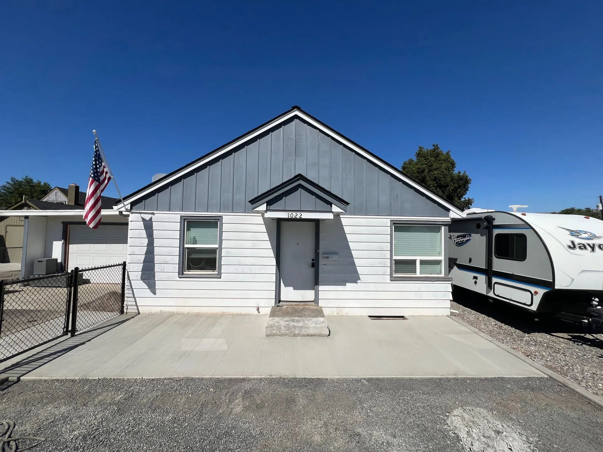 White house with blue roof, American flag, concrete pad, trailer parked to the right.