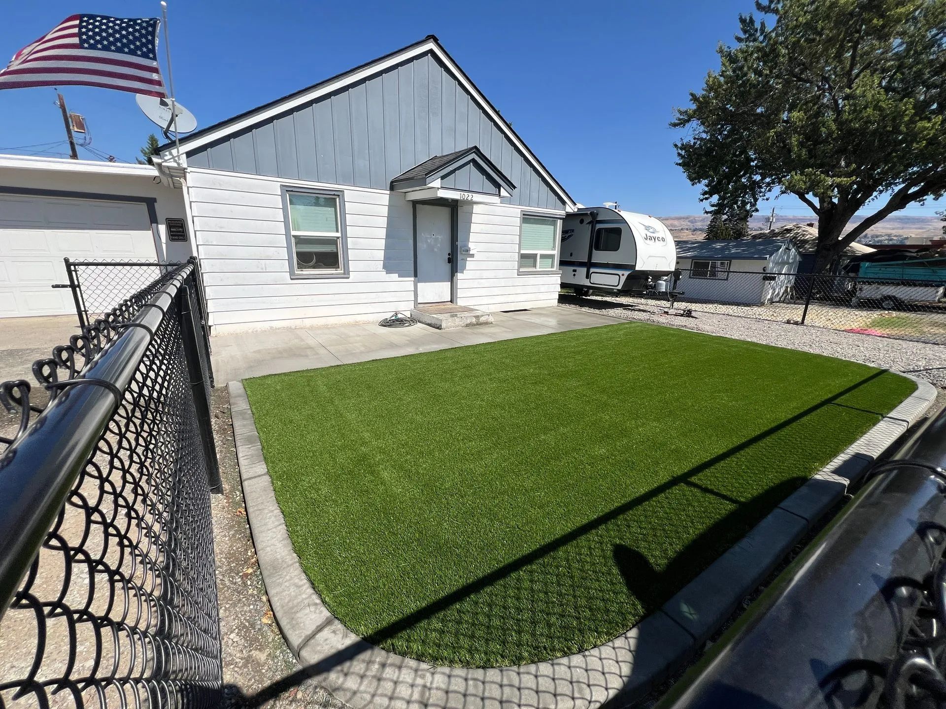 Small white house with a green artificial lawn, black chain-link fence, and an American flag.