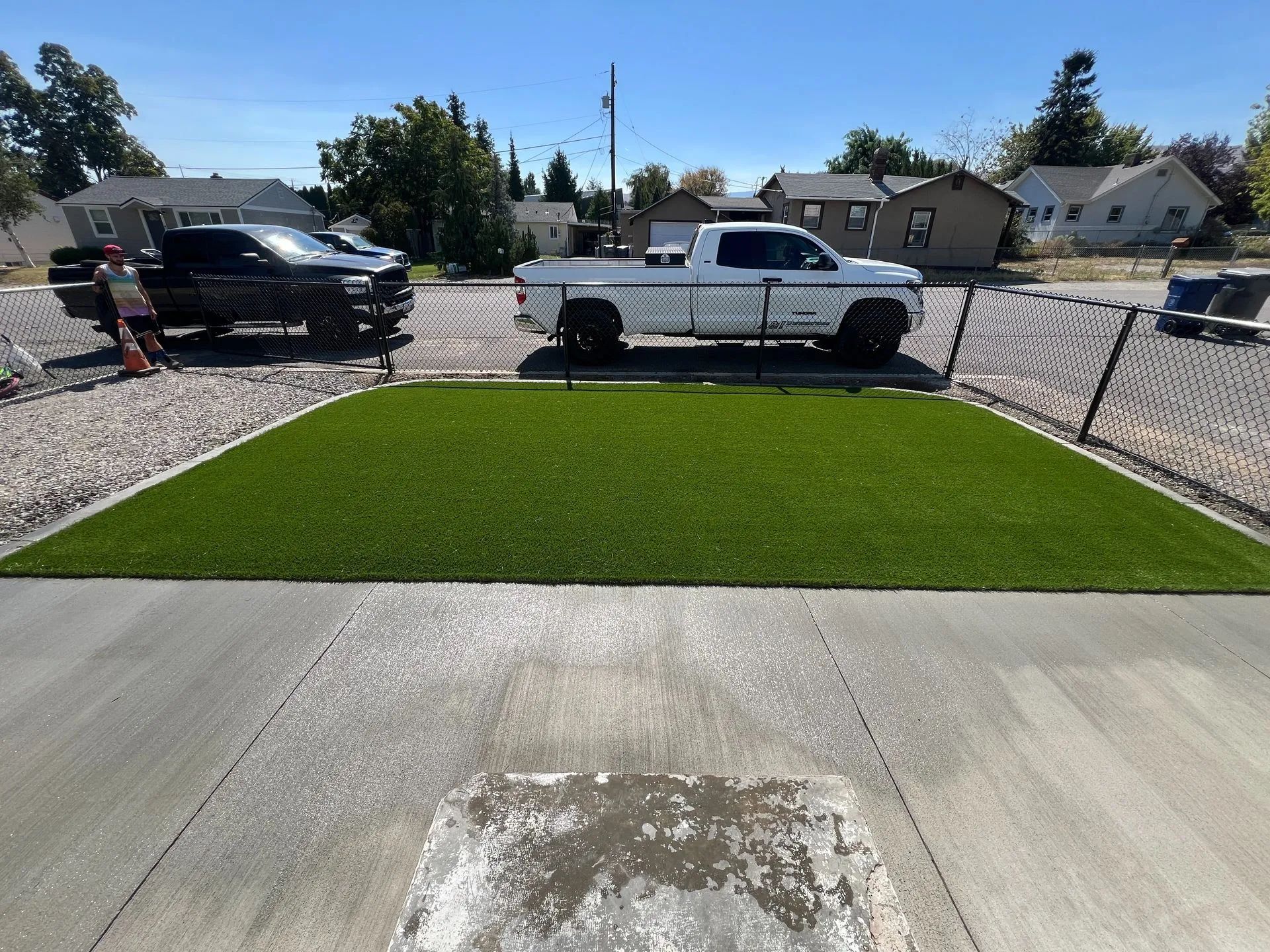 A patch of green artificial turf between concrete and two parked trucks on a sunny day.
