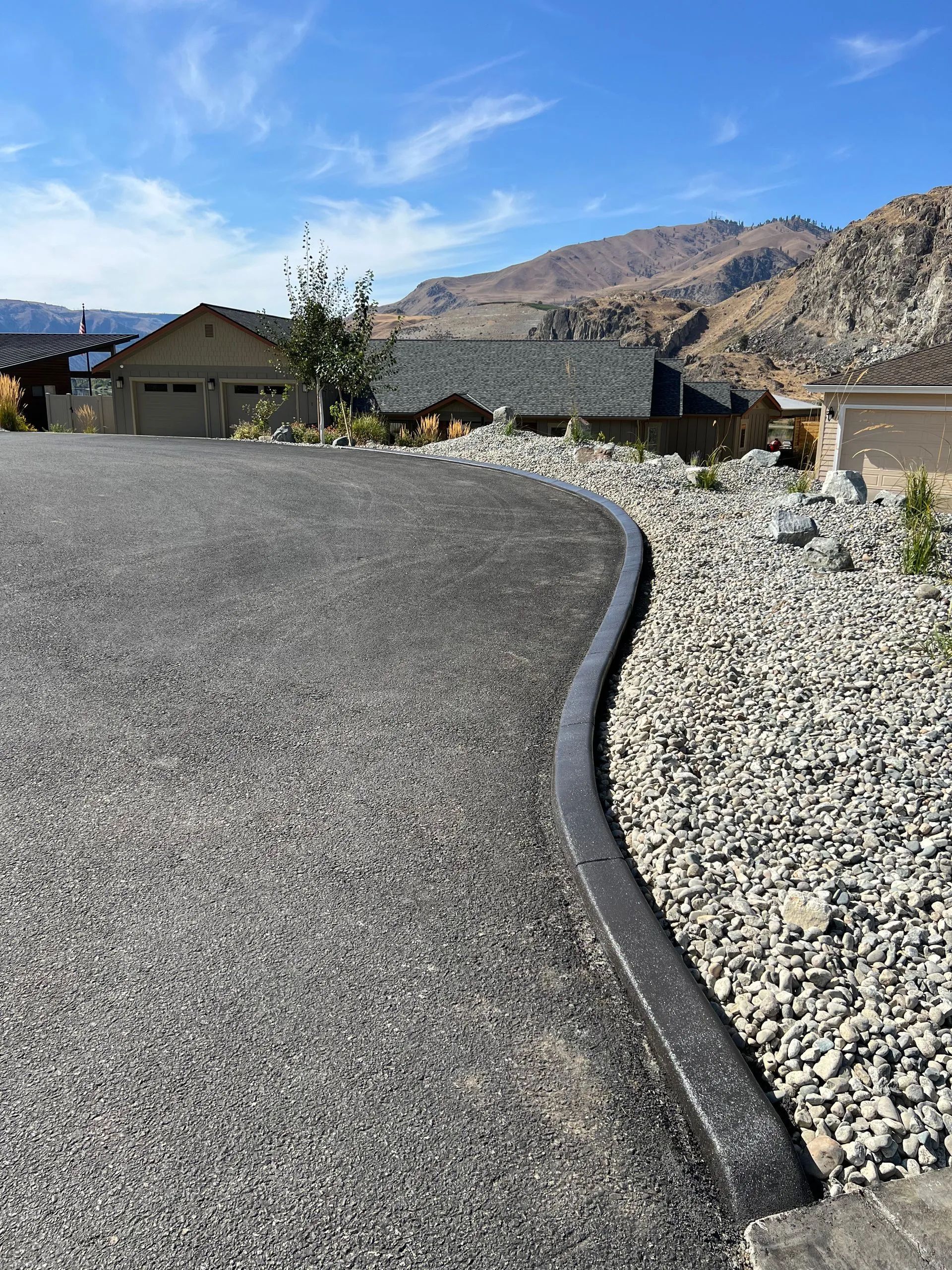 Asphalt driveway with dark edging borders a gravel bed in front of houses; mountains in background.