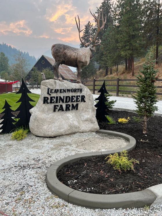 Sign for Leavenworth Reindeer Farm: stone, reindeer sculpture, black trees, gravel path, and greenery.