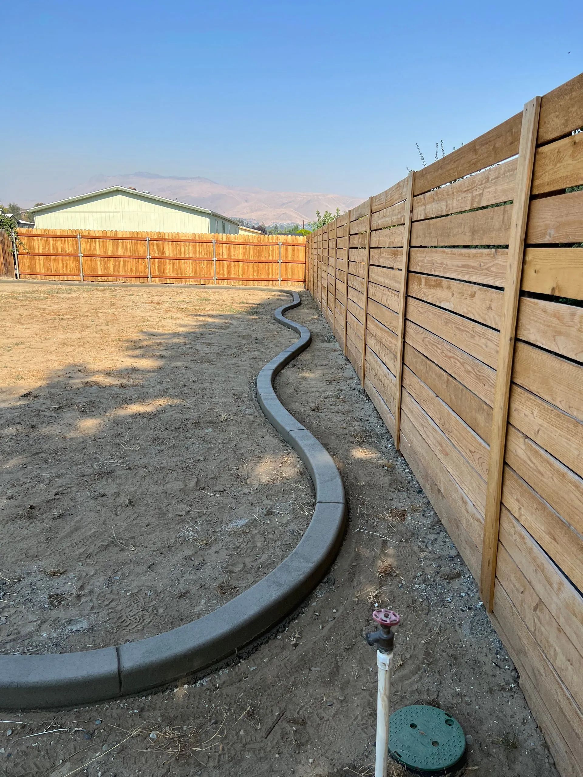 Curved gray concrete curb along a wooden fence in a yard with dirt and a water pipe.