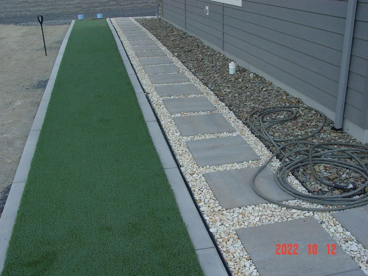 Green artificial turf path next to stepping stones and gravel bed against a house wall.