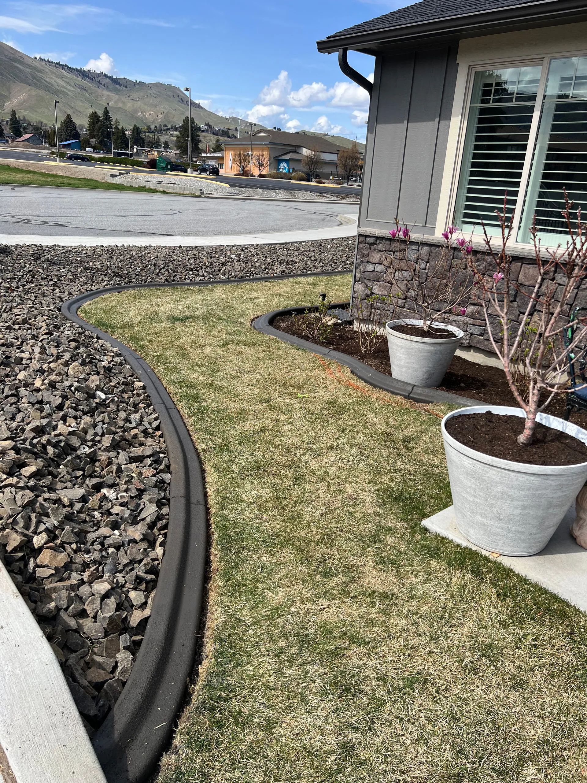 A curved grass pathway with dark edging and rock border, leading to a house with planters and shutters.