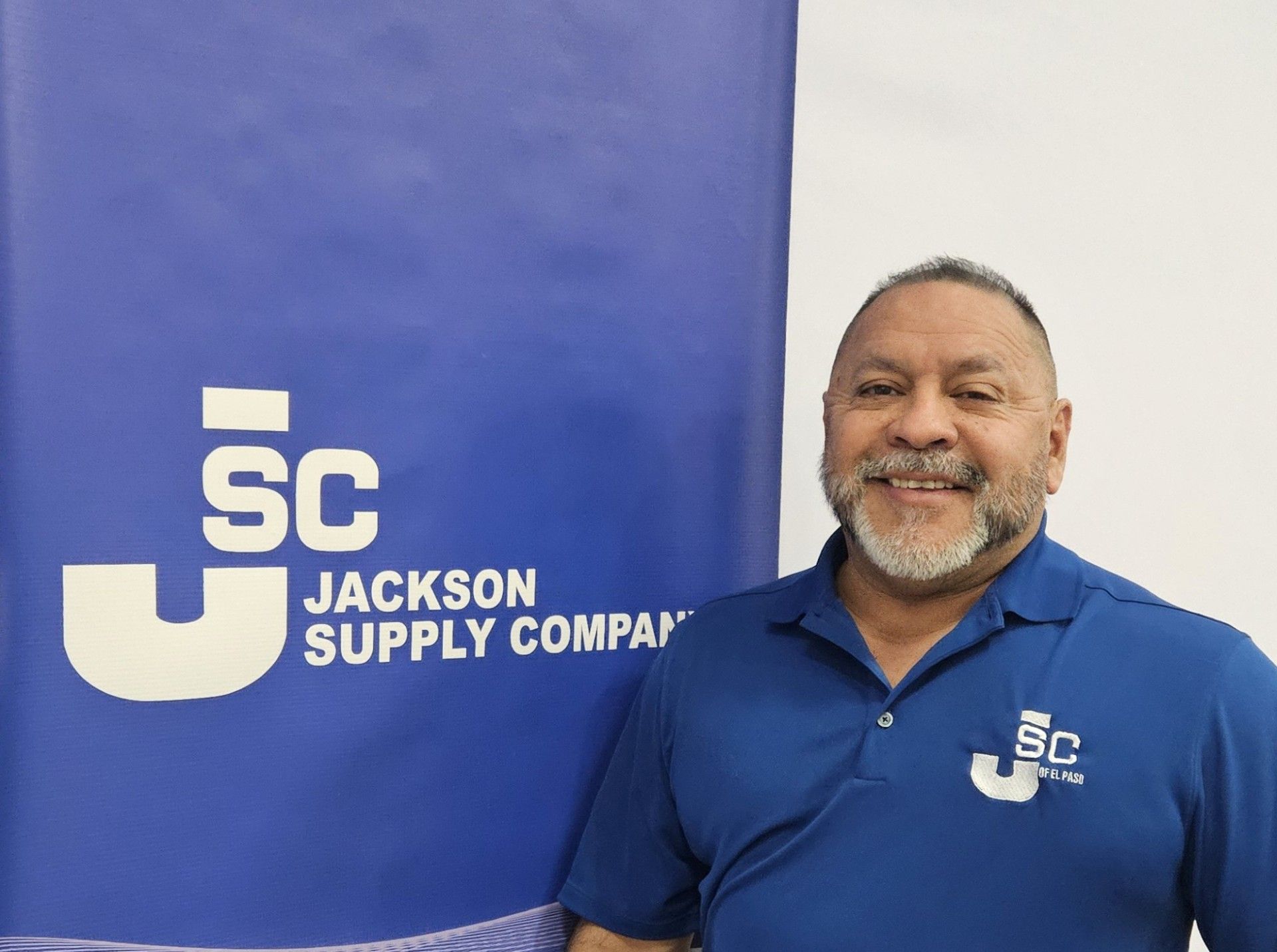 A person smiling in a blue polo shirt standing in front of a blue Jackson Supply Company logo banner.