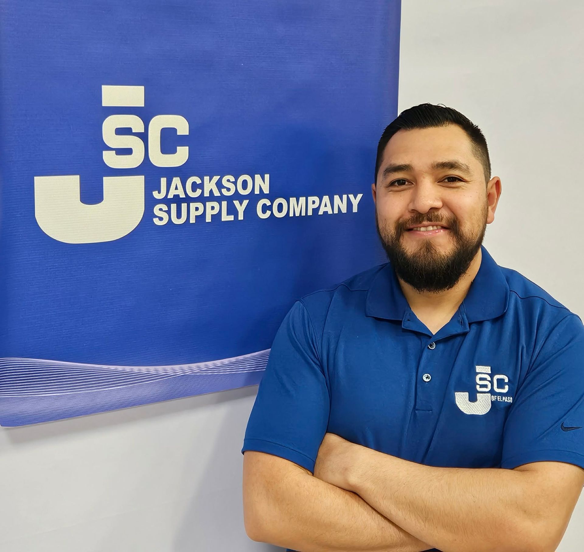 A smiling person in a blue polo shirt with a Jackson Supply Company logo stands before a matching branded backdrop.