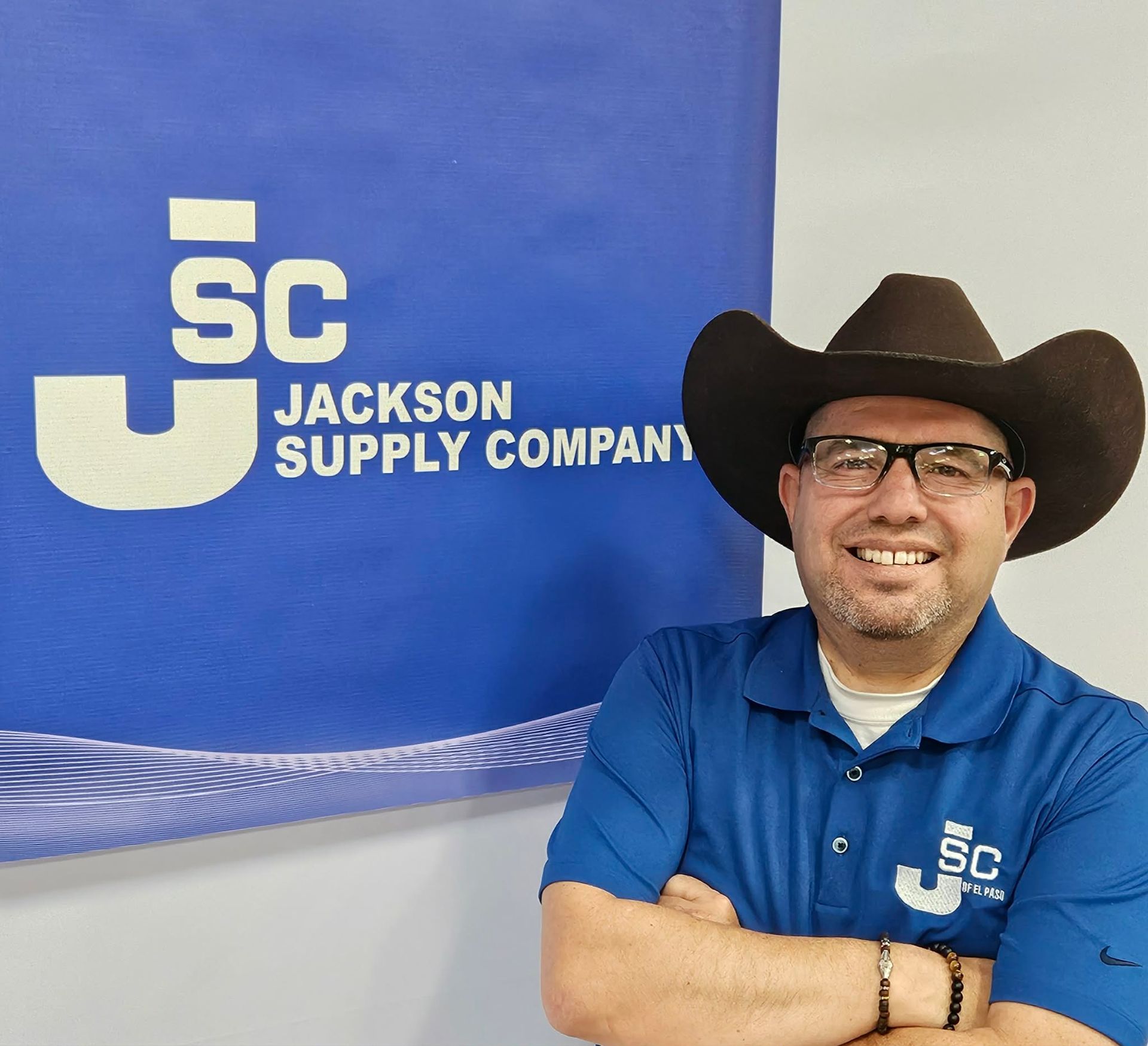 A person wearing a cowboy hat and a blue Jackson Supply Company shirt smiles with arms crossed in front of a company sign.