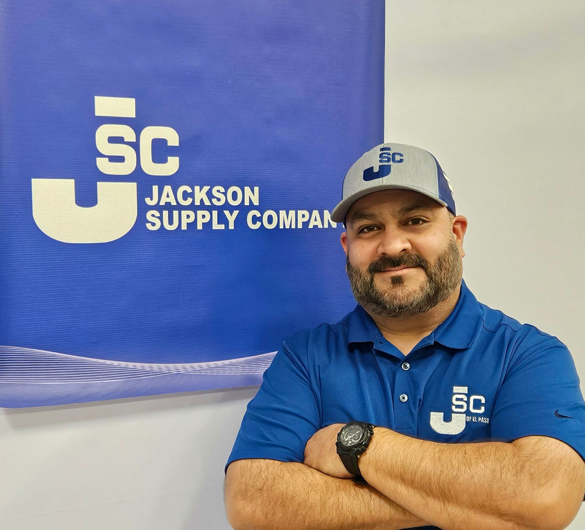 A person in a blue polo and matching cap with the Jackson Supply Company logo standing arms crossed before a branded sign.