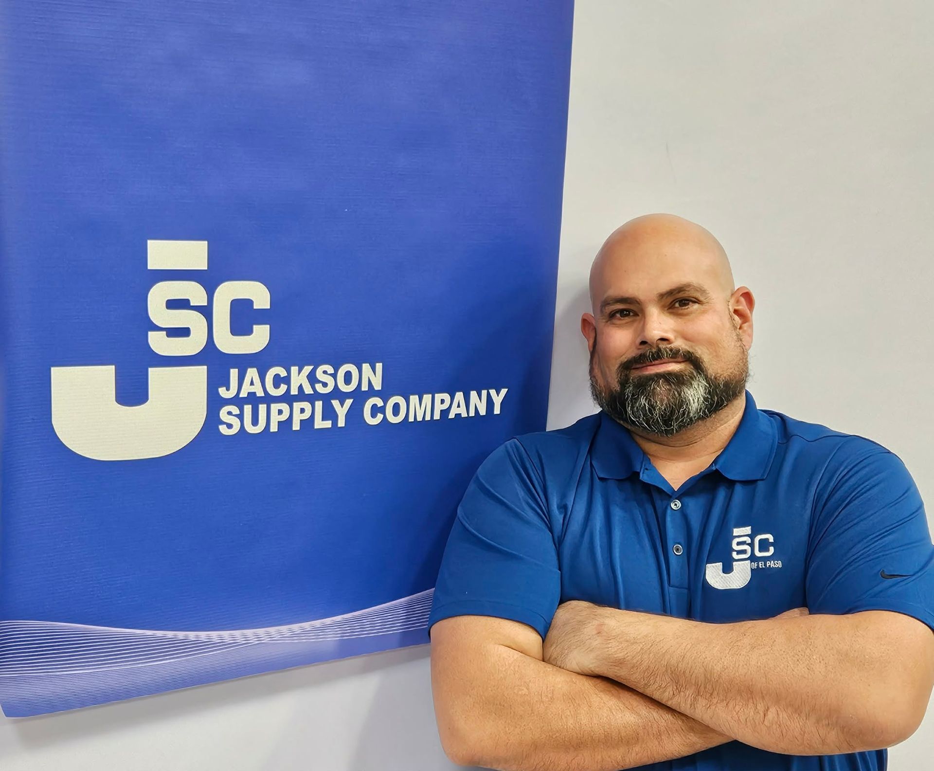 A man with a beard and a blue polo shirt standing with arms crossed next to a Jackson Supply Company blue and white banner.