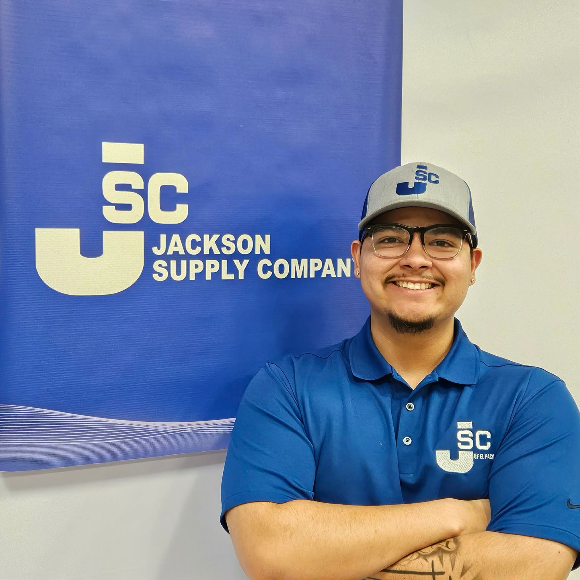 A smiling employee wearing a company polo and cap stands in front of a blue Jackson Supply Company logo banner.