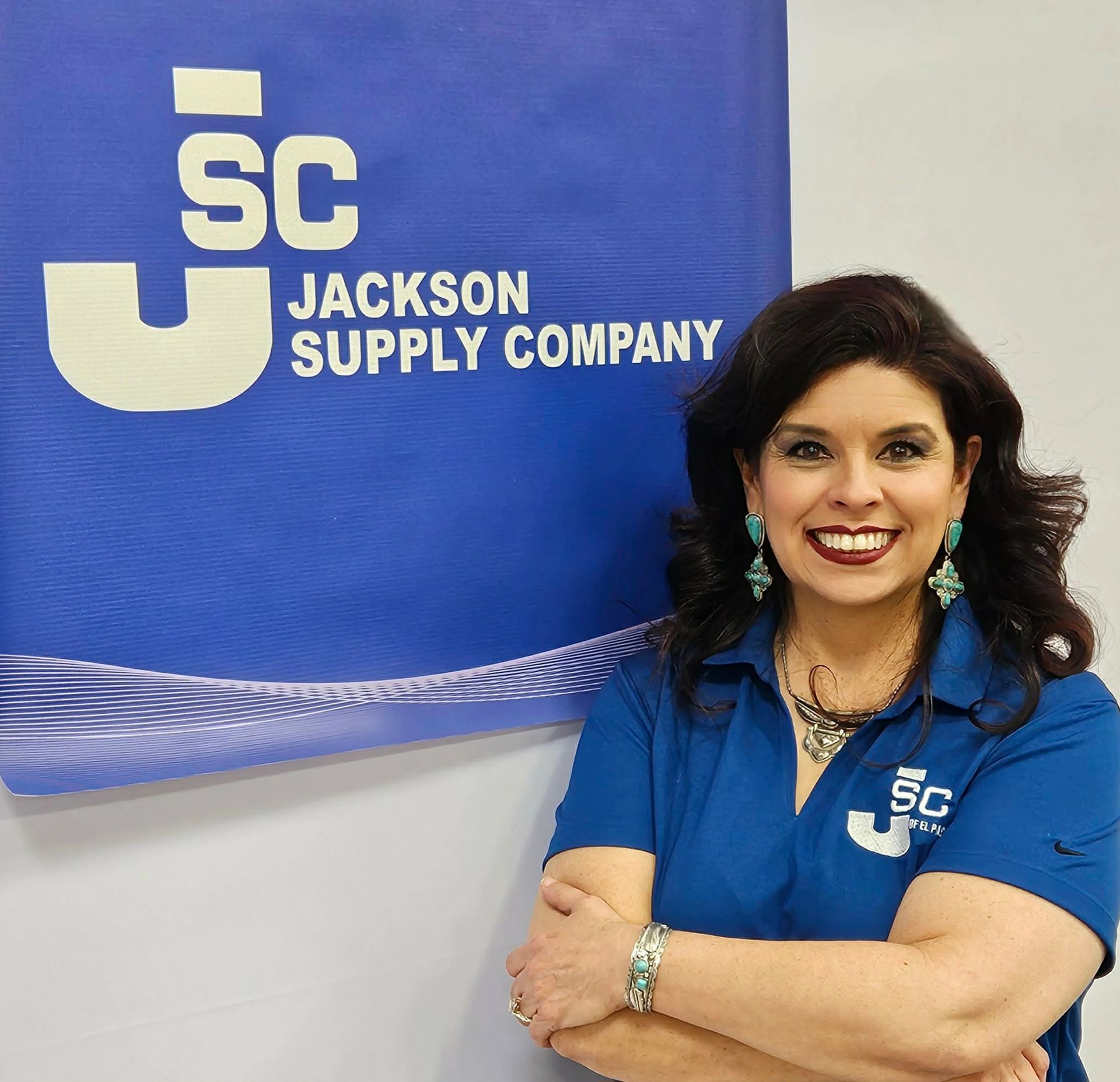 A smiling person wearing a blue polo shirt stands in front of a blue Jackson Supply Company logo sign.