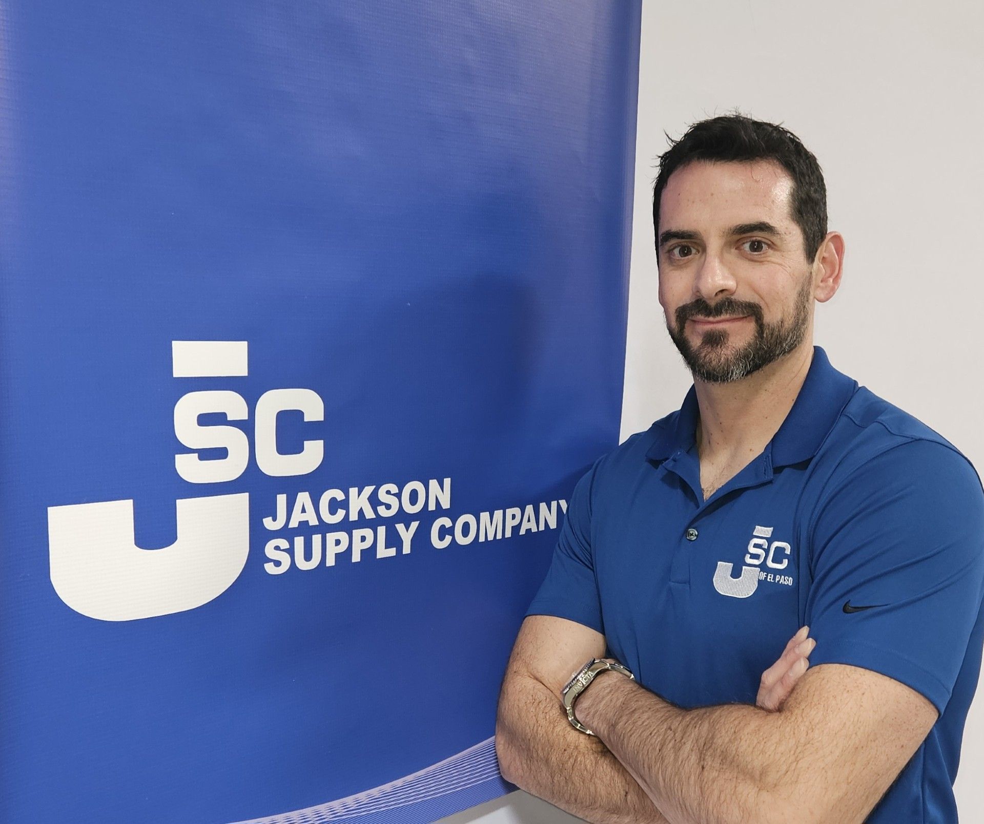 A man in a blue polo shirt with a Jackson Supply Company logo stands with arms crossed beside a matching blue company sign.