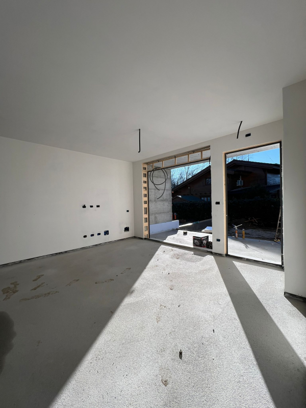 Stanza vuota e luminosa con pavimento in cemento grezzo e grandi porte finestre che si aprono su un patio.