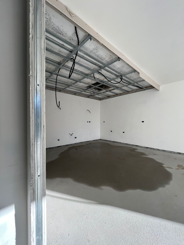 Stanza vuota e non rifinita con travi del soffitto a vista e una riparazione del pavimento in cemento bagnato.