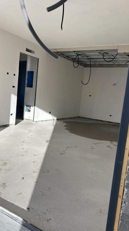 Stanza vuota e non rifinita con pavimento in cemento, telaio della porta blu e cavi elettrici a vista sul soffitto.