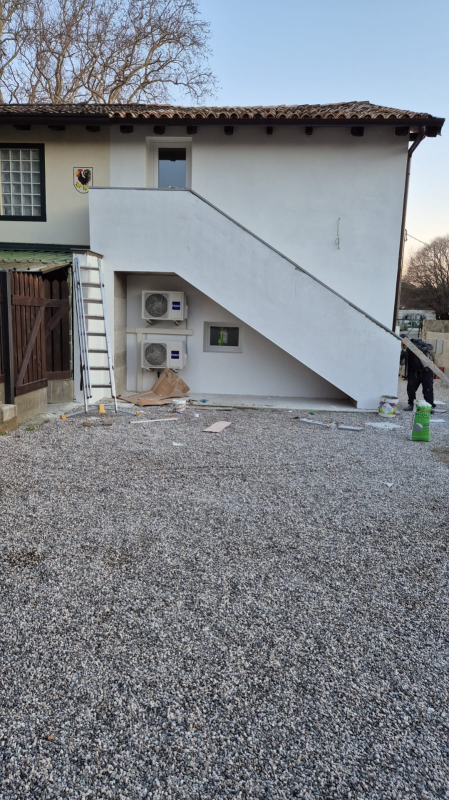 Esterno di un edificio bianco con scala diagonale, cortile ghiaioso e contatori delle utenze a parete.