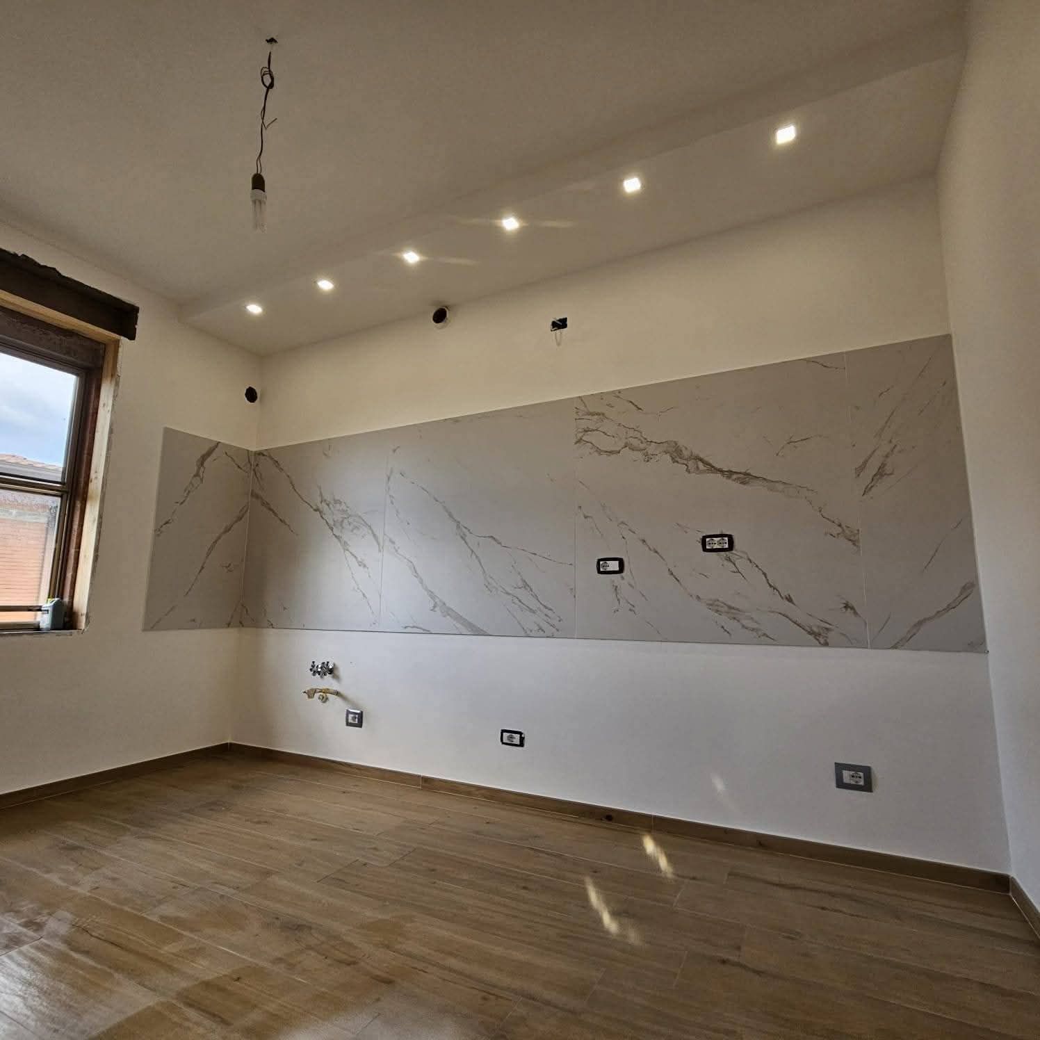 Stanza moderna vuota con pavimento in legno, pannello a parete effetto marmo, faretti a incasso e una finestra sulla sinistra.
