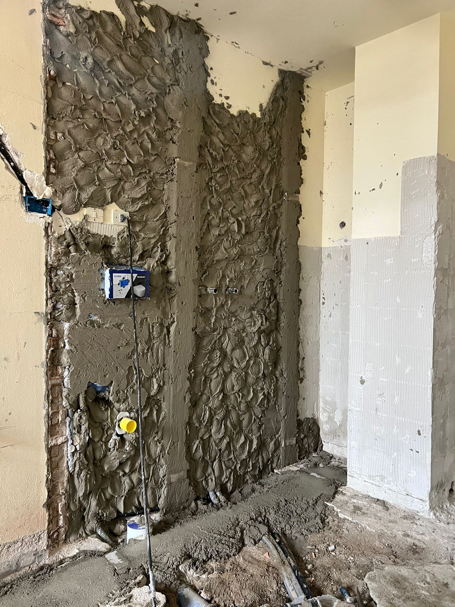 Parete del bagno non rifinita con intonaco grigio a vista, scarichi idraulici e detriti sul pavimento.