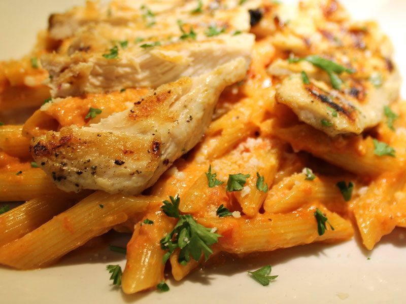 Grilled chicken breast over penne pasta in creamy tomato sauce, garnished with parsley.