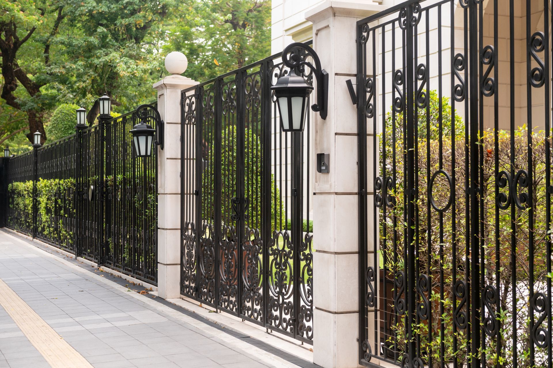 Black wrought-iron fence with stone pillars along a tree-lined sidewalk