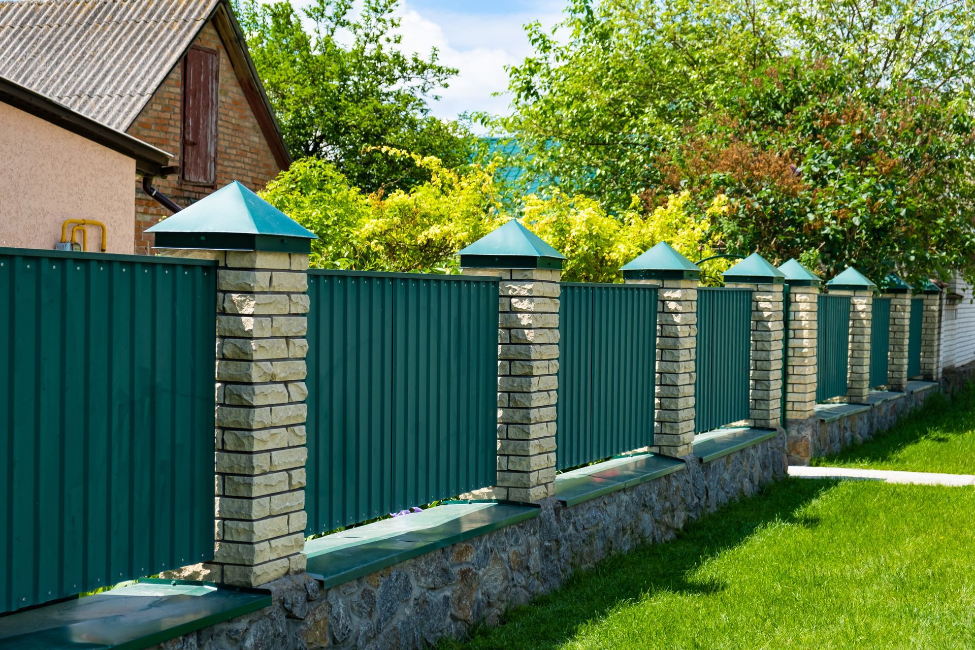 Green metal fence with stone pillars along a grassy yard beside a house and trees
