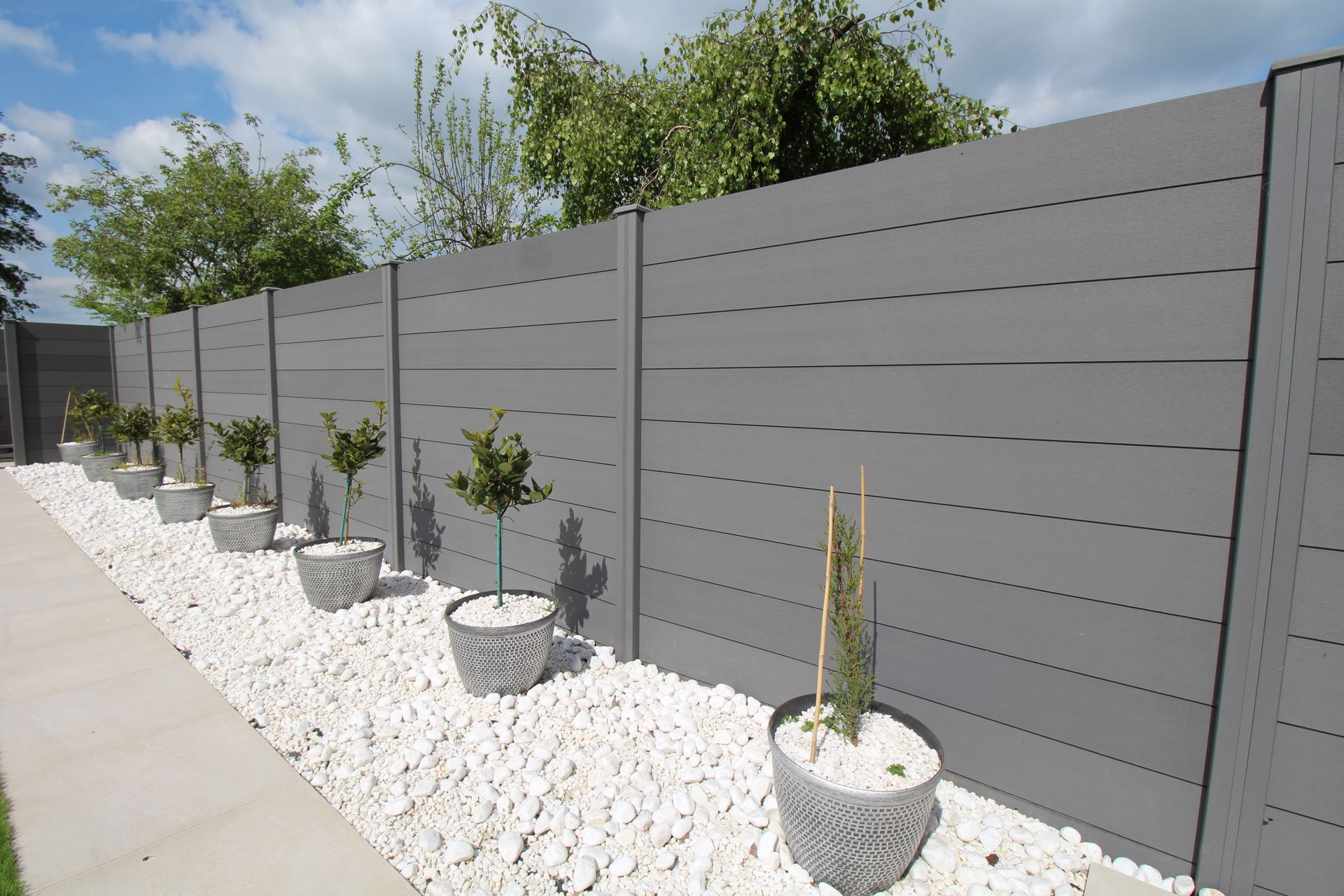 Gray slatted fence with white gravel and potted plants along a sidewalk under a blue sky