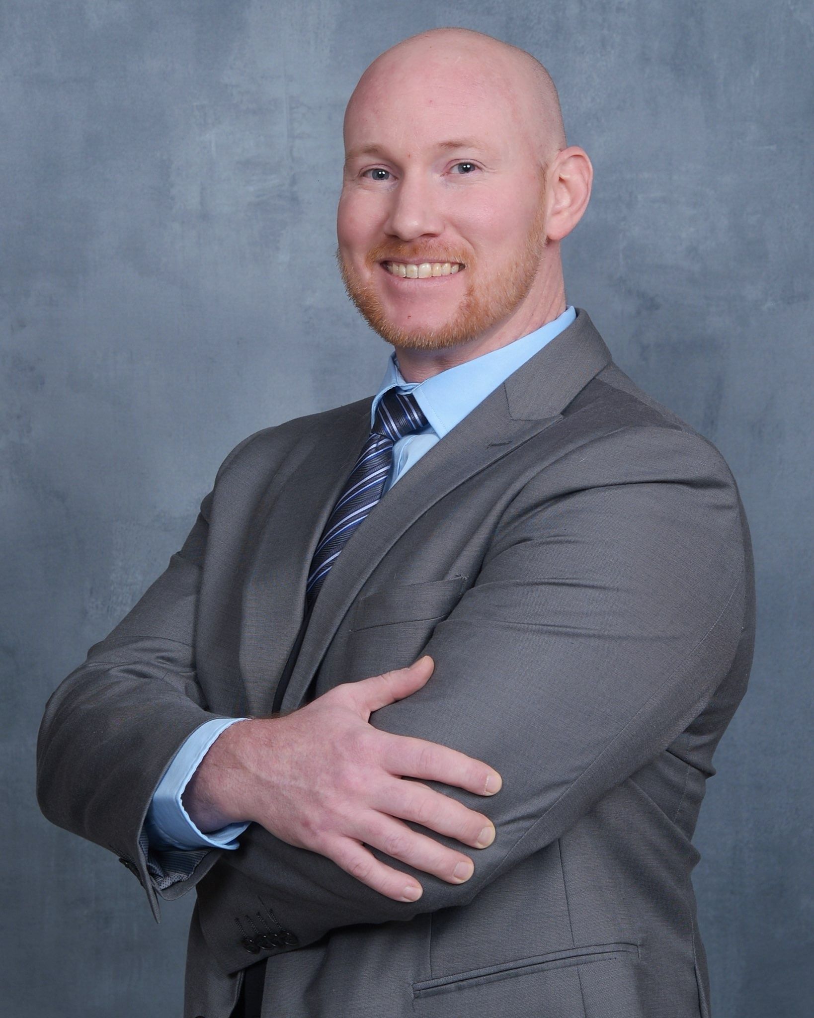 A smiling person with a shaved head wears a gray suit, light blue shirt, and patterned tie, posing with arms crossed.