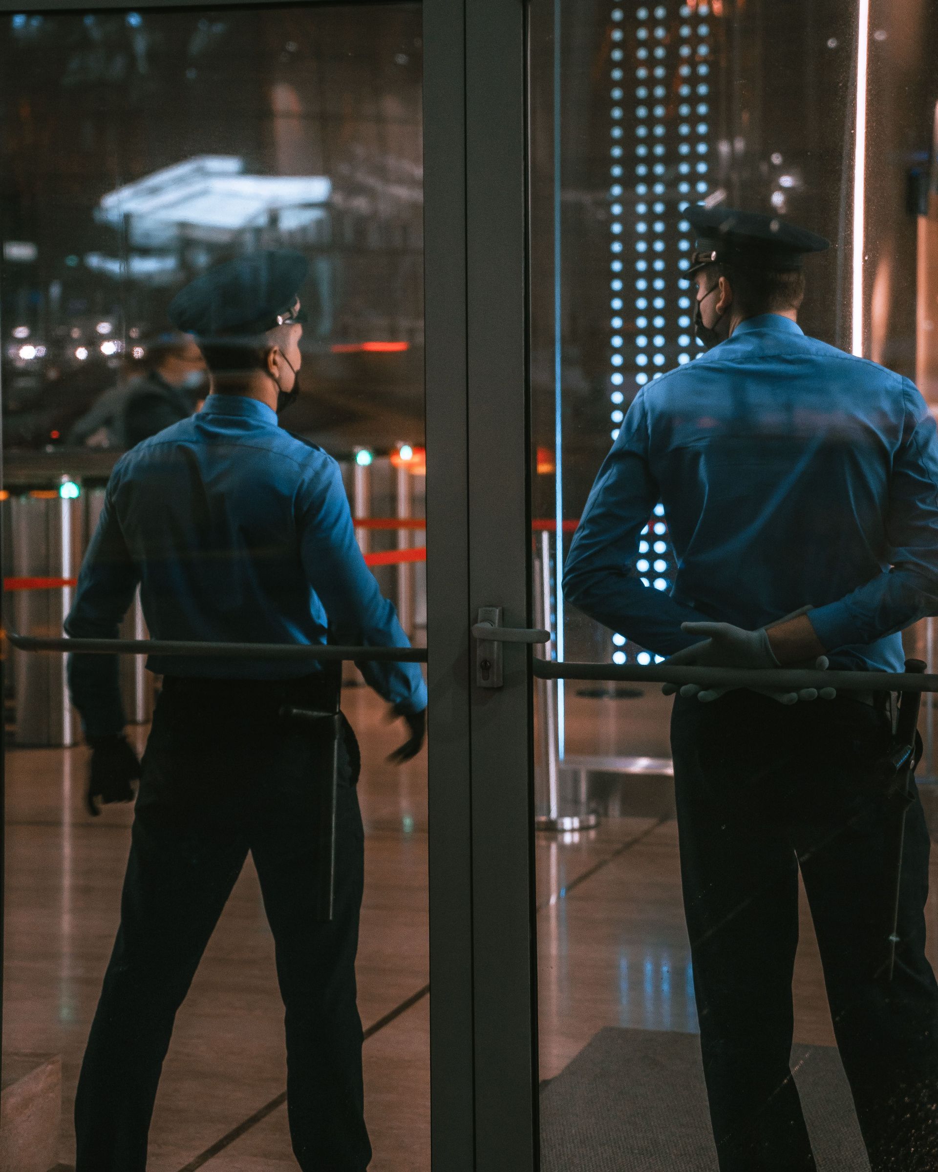 Two security guards stand at a glass entrance at night, city lights reflected behind them.