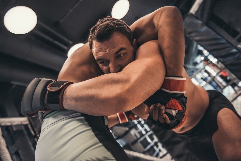 Two athletes grappling in a mixed martial arts gym, one in a clinch with gloves and wraps