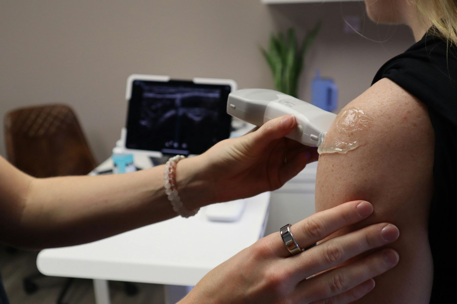 Person receiving an ultrasound on their arm. Technician holds device with gel. Digital display in background.