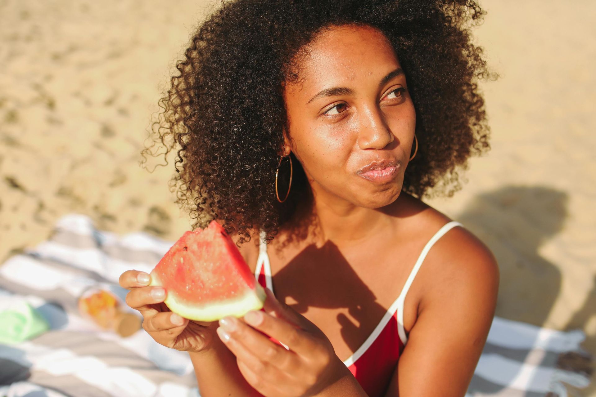 Woman eating watermelon on beach; wearing red swimsuit, enjoying summer sun.