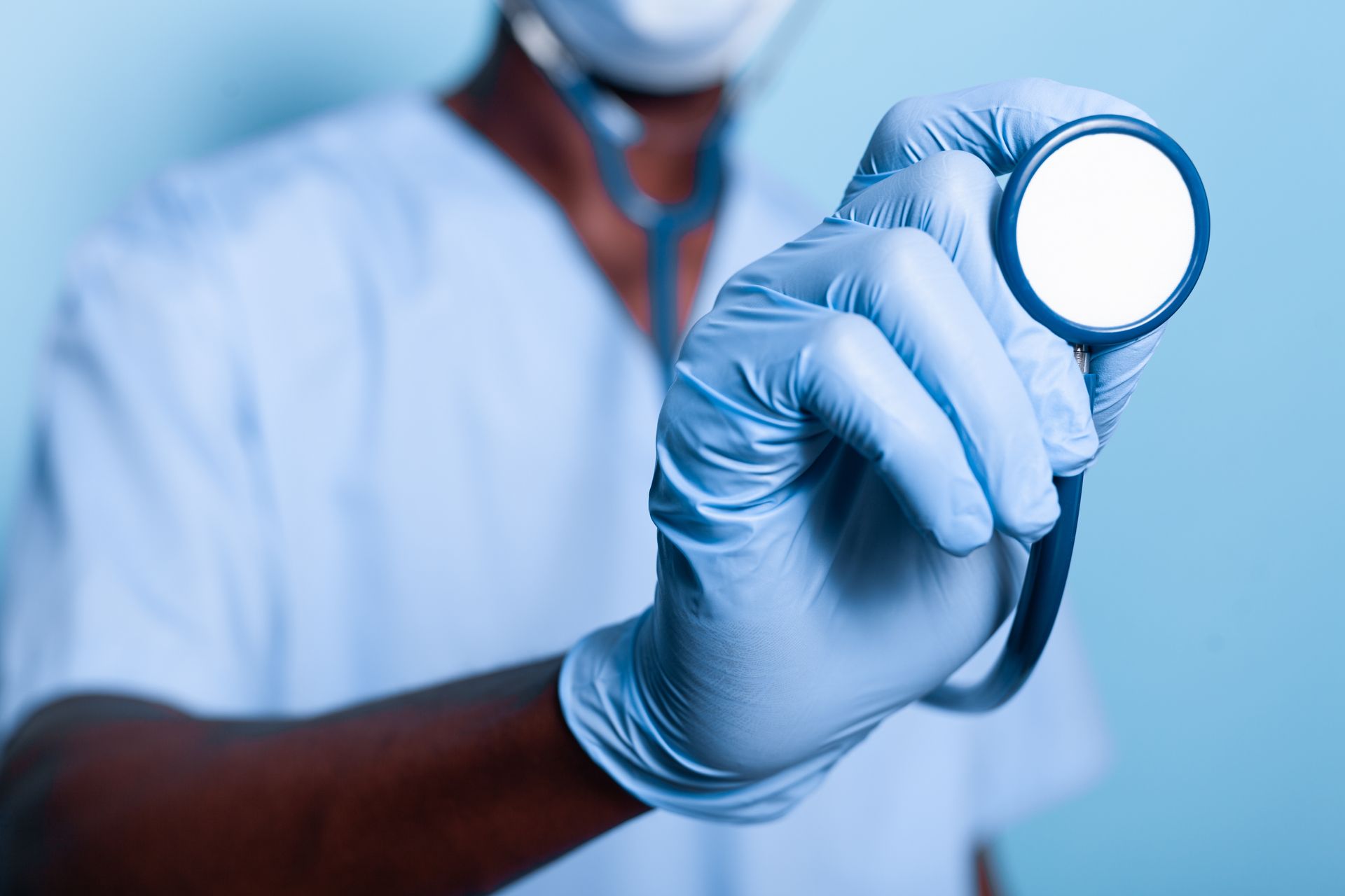 A doctor wearing a mask and gloves is holding a stethoscope in his hand.