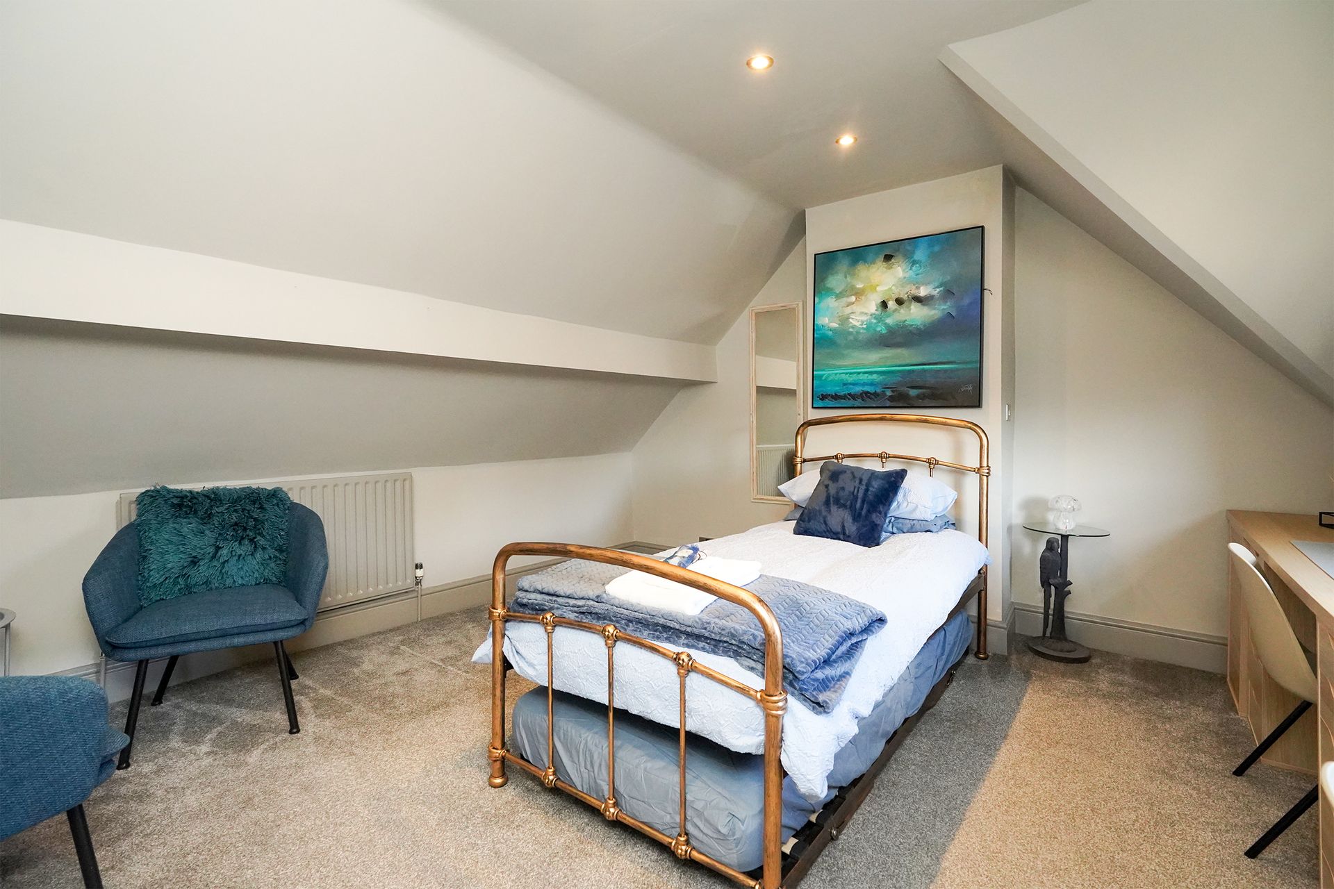 Small attic bedroom with twin bed, blue chair, desk, and sloped white ceiling.