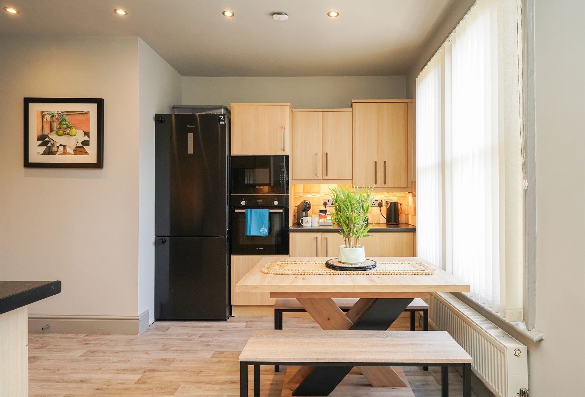 Modern kitchen with light wood cabinets, black refrigerator, and dining table by a bright window.