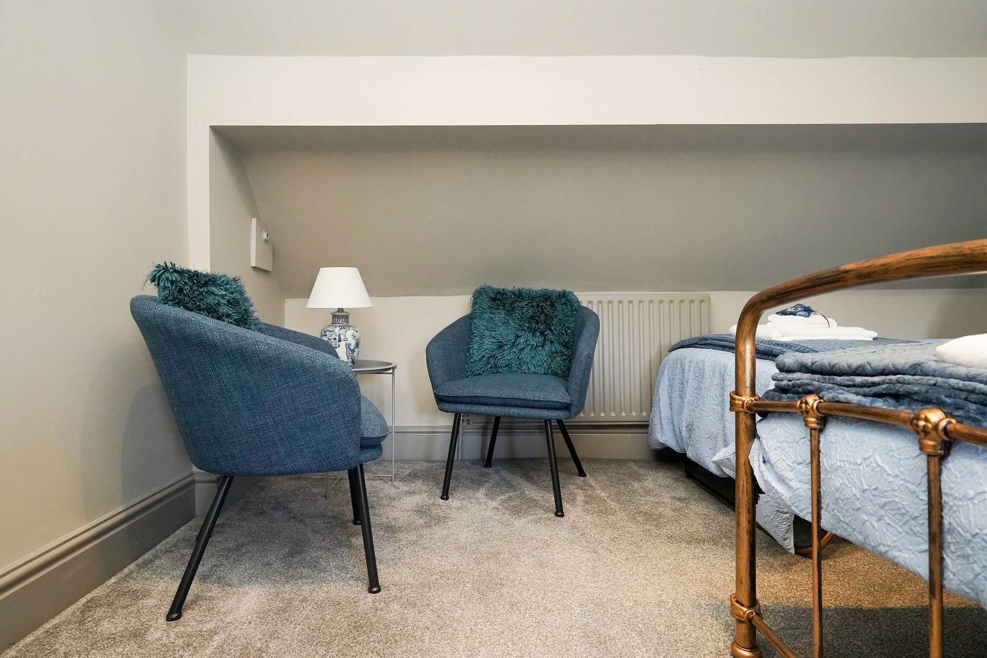 Small bedroom with blue chairs, lamp, bed, and wicker bed frame in a cozy attic-like room