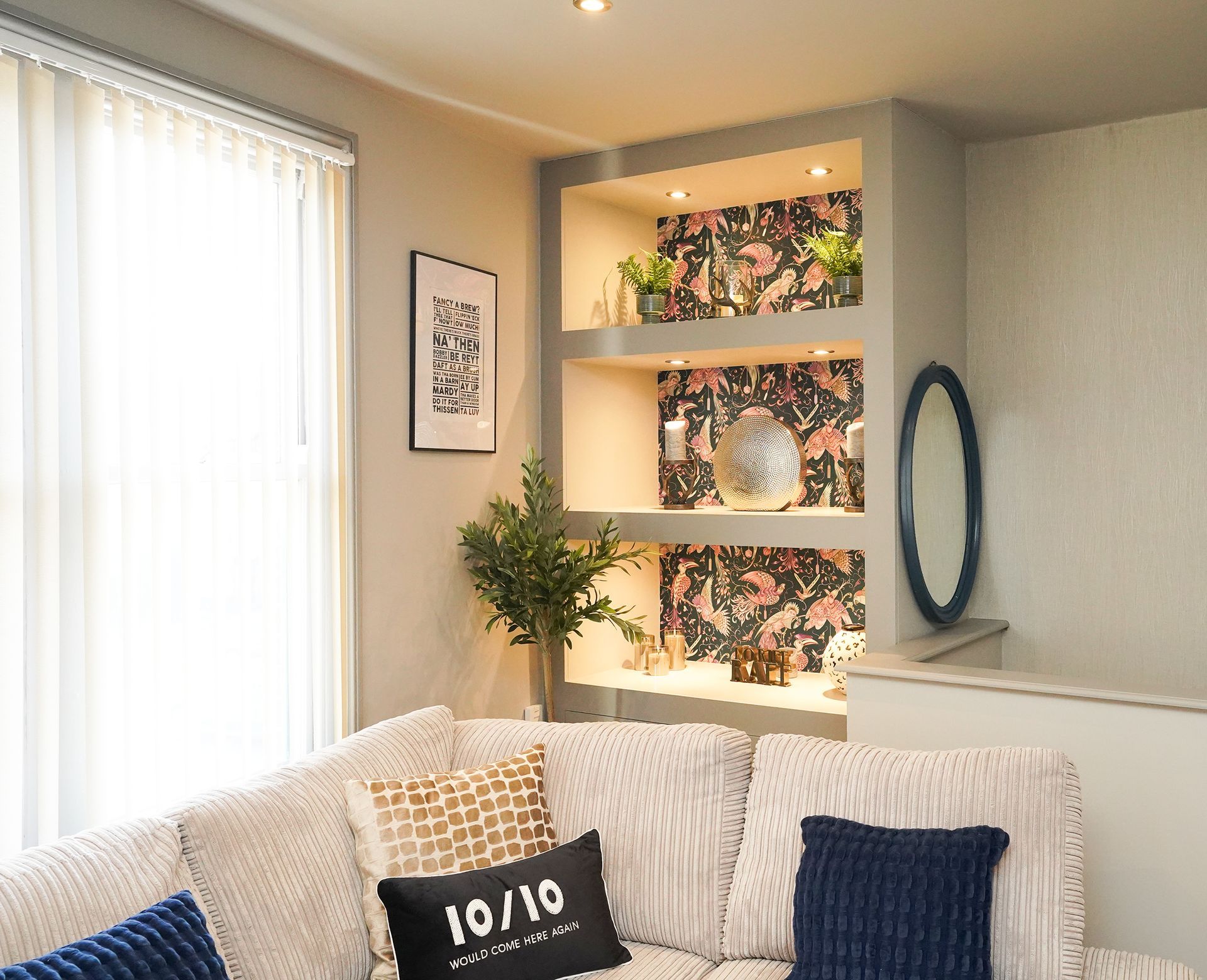 Bright living room with sofa, blue pillows, wall art, shelves, and a window with sheer curtains