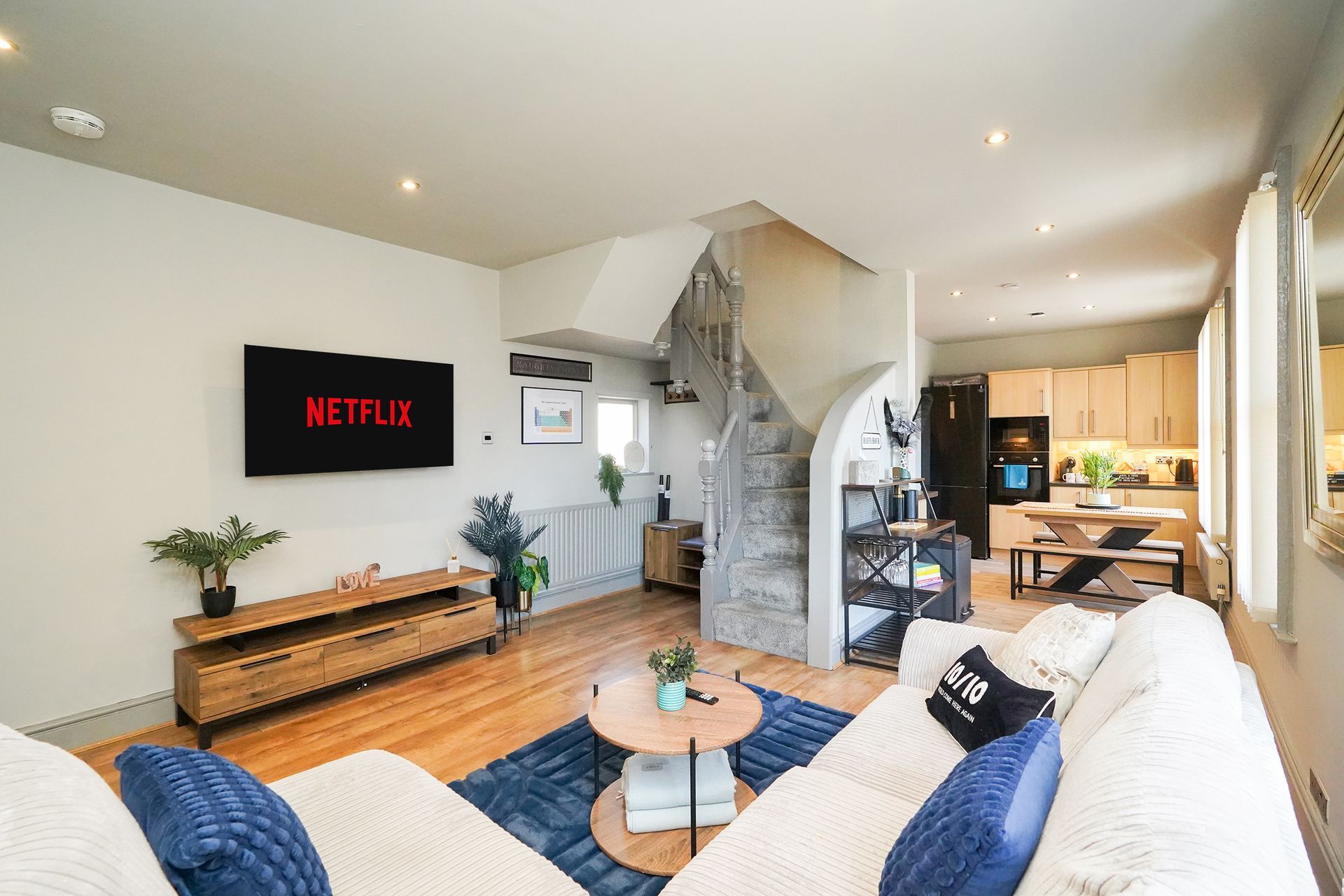 Bright living room with white sofa, wood floor, TV, staircase, and dining area in back